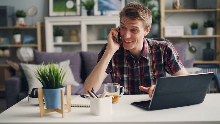 Hombre joven con genes buenos, sonriendo mientras habla por teléfono y usa una laptop en su hogar moderno.