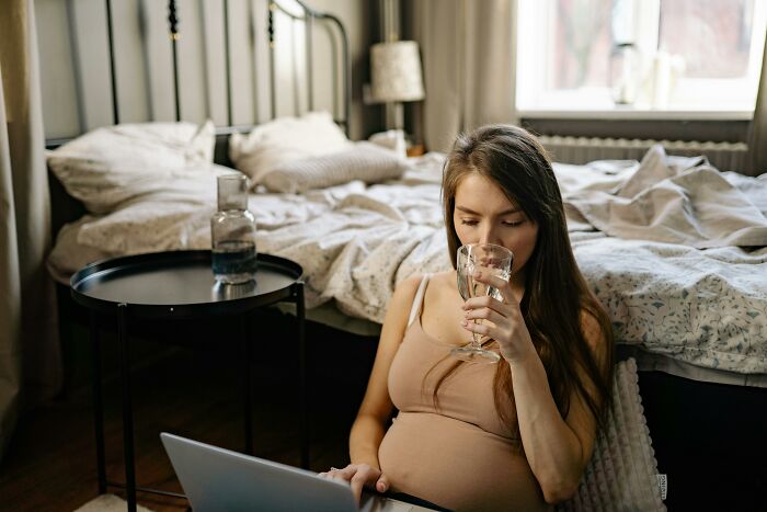Mujer embarazada escuchando conversaciones privadas terroríficas mientras usa una laptop en una habitación con cama desordenada.