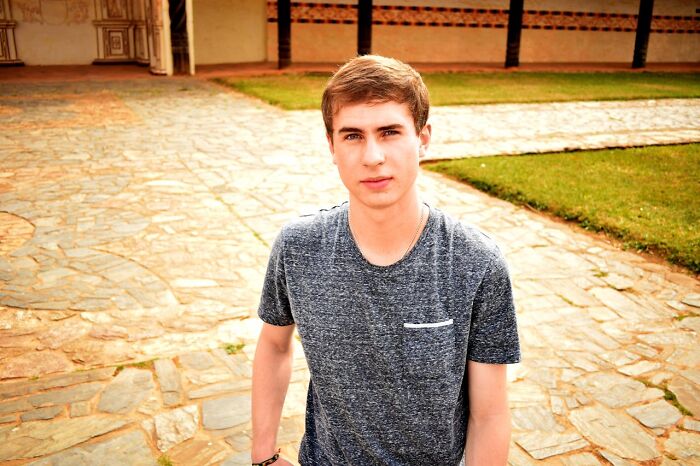 Joven de cabello claro y camiseta gris posando al aire libre en un camino de piedra, genes muy buenos o muy malos.