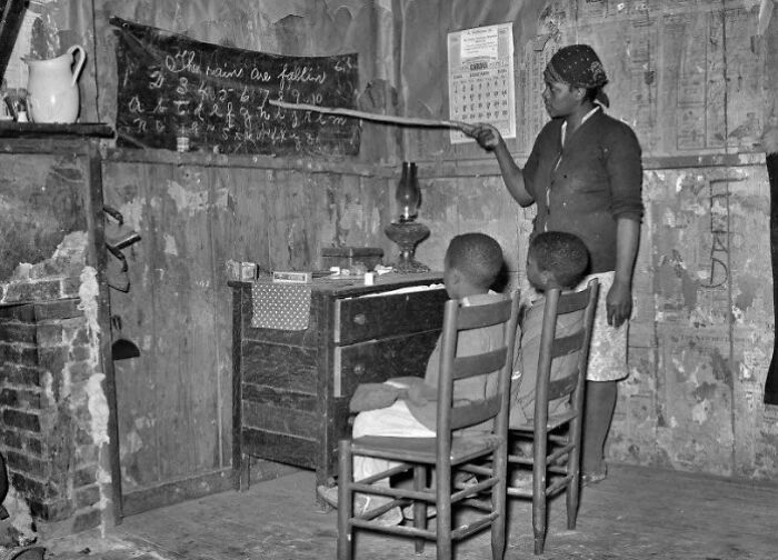 Clase en casa antigua con mujer enseñando a dos niños, mostrando un lado distinto de la historia en una foto histórica en blanco y negro.