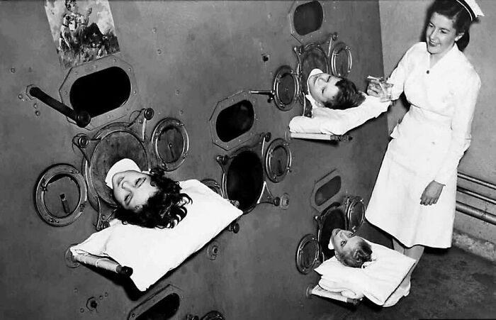 Mujer enfermera atendiendo a tres personas en cámara de aislamiento, mostrando un lado distinto de la historia médica.
