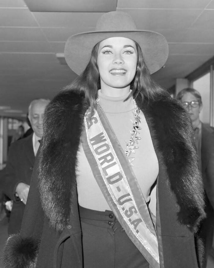Mujer vestida con abrigo de piel y sombrero, con banda de concurso, mostrando un lado distinto de la historia.