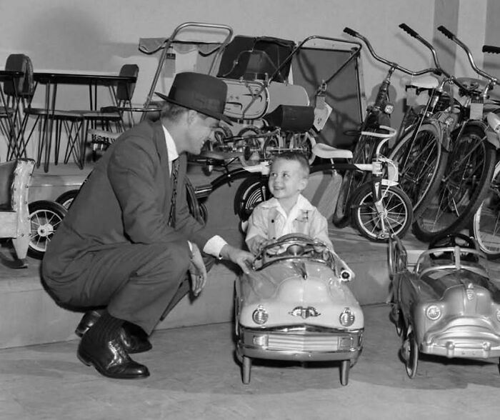 Hombre con sombrero junto a niño en coche de juguete antiguo, mostrando un lado distinto de la historia familiar.
