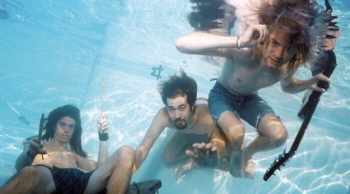 Tres jóvenes con guitarras sumergidos en una piscina, imagen con ángulos alternativos y perspectiva original.