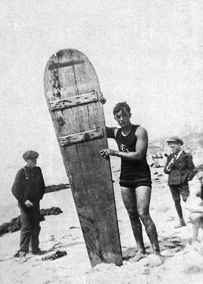 Joven con curiosa tabla de surf antigua en la playa, mostrando fotos históricas del pasado inédito en blanco y negro.