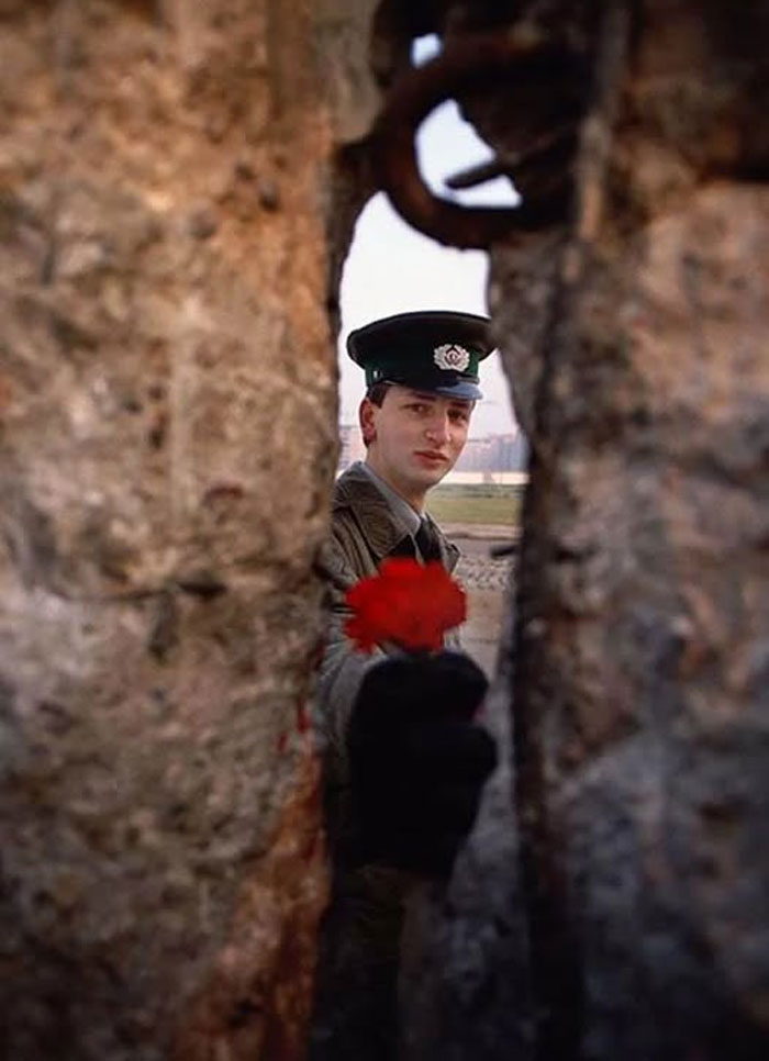 Soldado de frontera con gorra y guantes ofreciendo una flor roja entre muros antiguos en foto histórica curiosa del pasado.