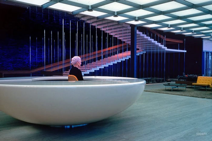 Mujer mayor sentada en silla moderna blanca en interior minimalista con escalera flotante y muebles vintage, foto histórica curiosa.
