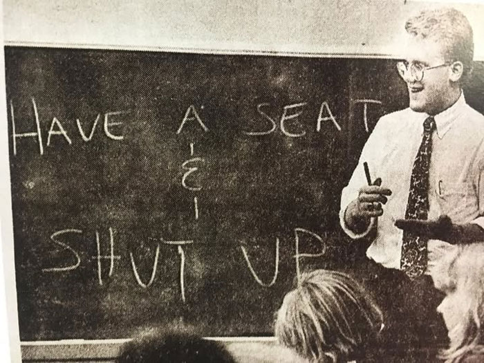 Profesor en aula con pizarra mostrando curiosa foto histórica que refleja métodos de enseñanza poco convencionales.