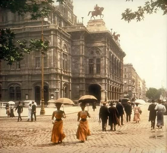 Personas con paraguas caminando frente a edificio histórico antiguo, clásica foto histórica del pasado inédito.