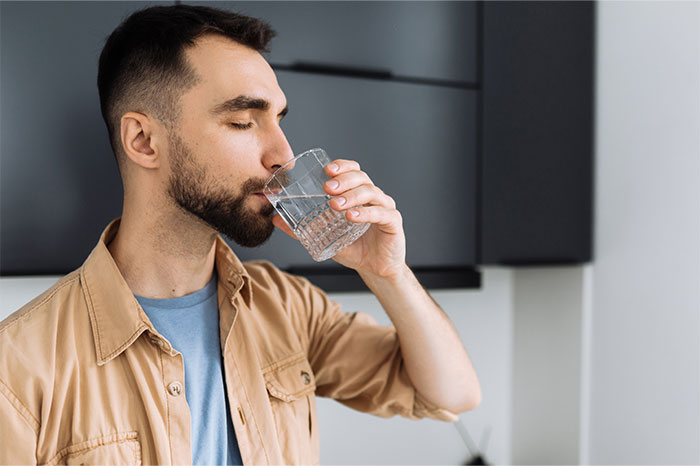 Hombre joven con barba bebiendo agua en su cocina, ilustrando la pérdida de atracción en parejas según mujeres.