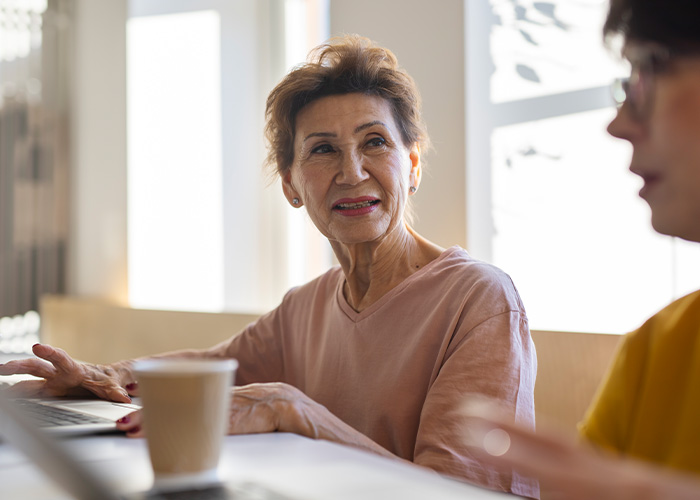 Mujer mayor conversando con otra persona en cafetería, ilustrando experiencias que soportan con sus suegras.