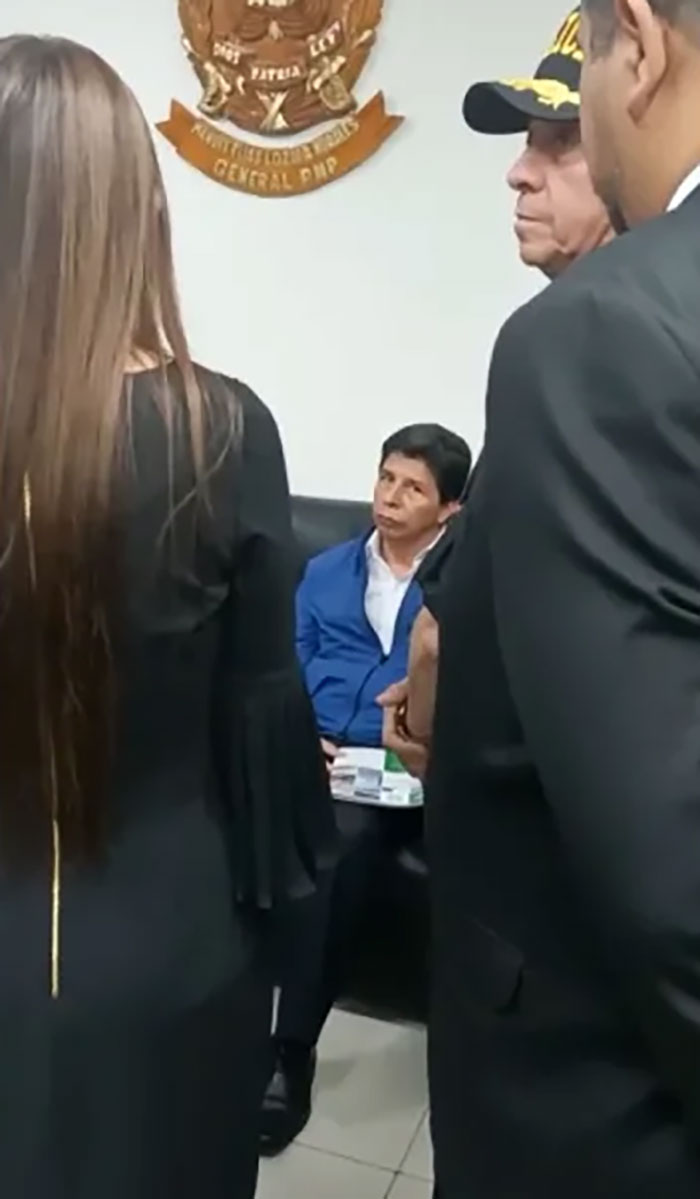 Hombre sentado con chaqueta azul rodeado por personas en traje formal, en contexto laboral con ambiente serio y oficial.