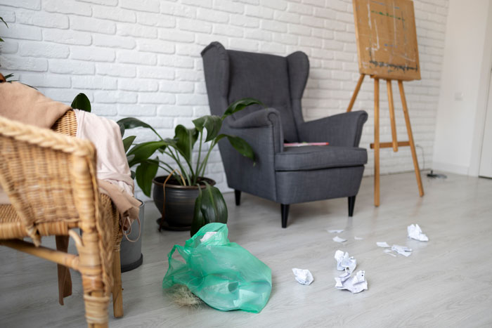 Sala con silla gris, planta, basura en bolsa verde y papeles arrugados en el suelo, ilustrando tendencias hogareñas superadas.