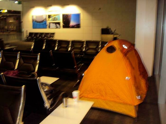 Tienda de campaña naranja instalada en sala de espera de aeropuerto, mostrando actitud de personas a las que todo les da igual.