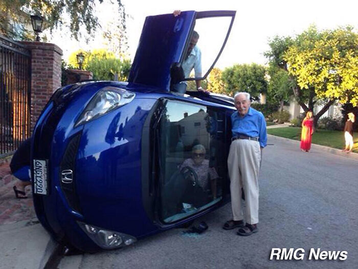 Hombre mayor tranquilo junto a coche volcado en calle residencial reflejando actitud de personas a las que todo les da igual.