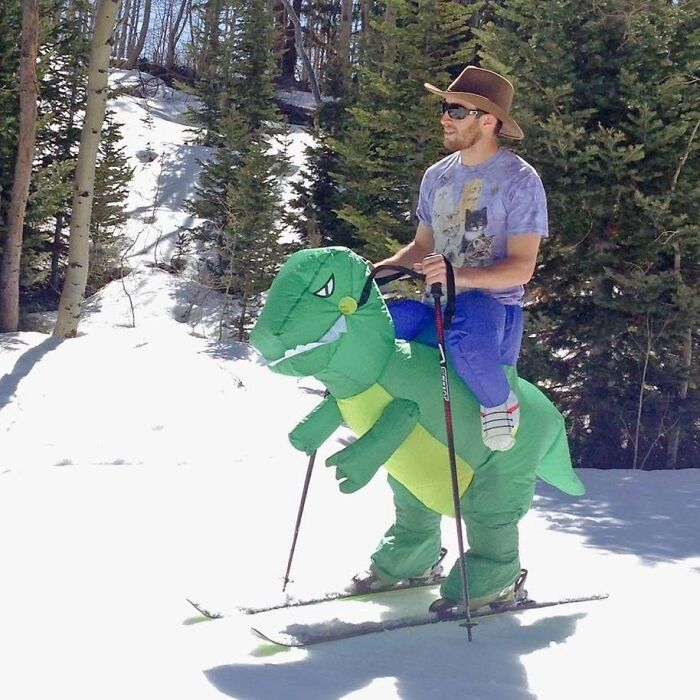 Hombre con disfraz de dinosaurio esquiando en la nieve, mostrando actitud relajada y despreocupada.