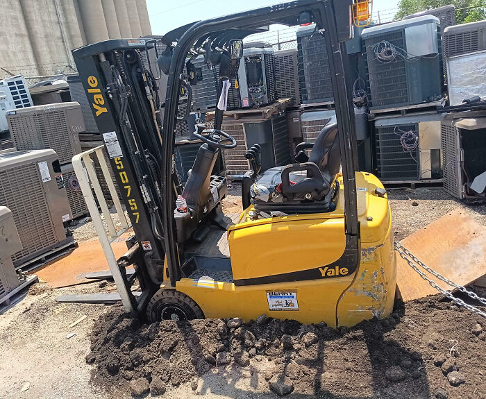 Montacargas amarillo atascado en el barro en un lugar de trabajo con varios equipos industriales alrededor.