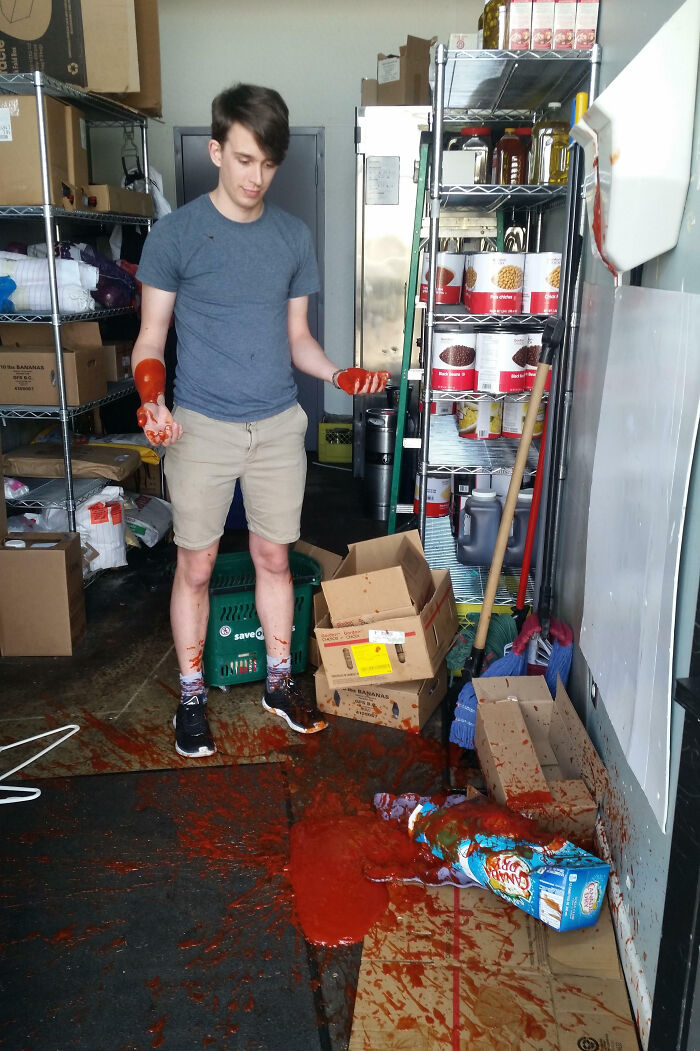 Joven con camiseta gris y shorts en almacén con salsa derramada y cajas desordenadas, día peor en el trabajo.