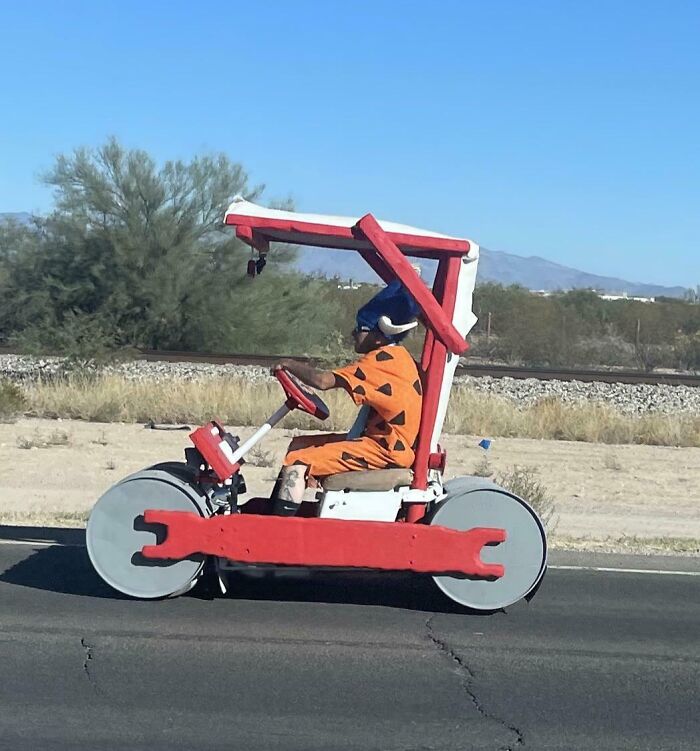 Hombre vestido como Pedro Picapiedra conduciendo vehículo casero en carretera desierta, actitud despreocupada y original.