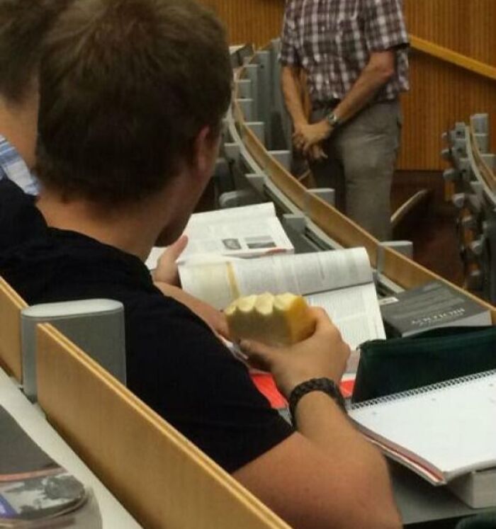 Estudiante en aula sosteniendo un pan sobre libros abiertos sin prestar atención, mostrando actitud de personas a las que todo les da igual.