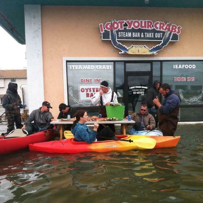 Personas a las que todo les da igual comiendo mariscos en kayak durante una inundación frente a un restaurante.