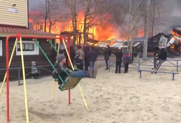 Persona en columpio ignorando incendio intenso en zona residencial con varias personas observando desde lejos.