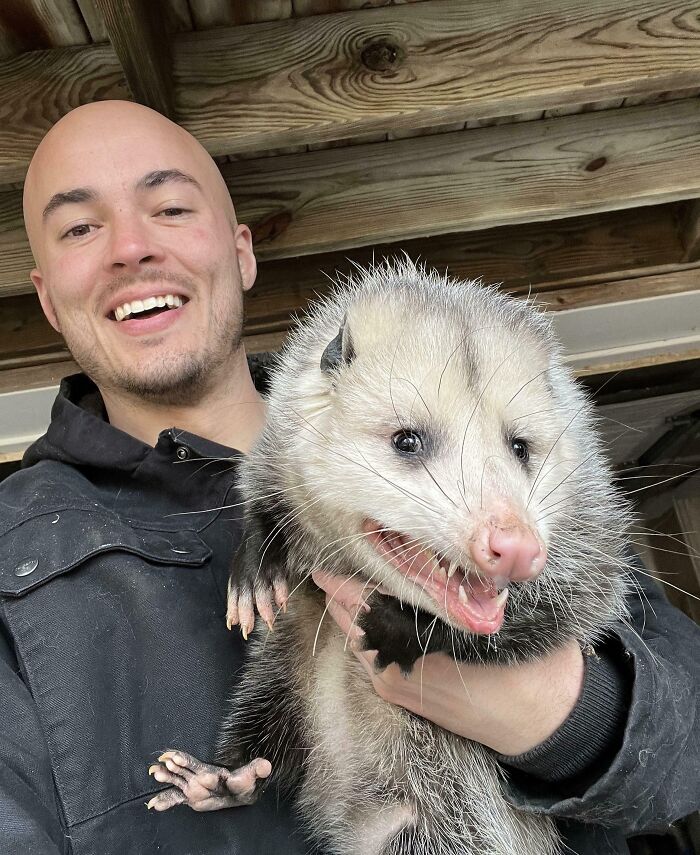 Hombre sonriente sosteniendo un animal divertido y absurdo que parece un zarigüeya en ambiente natural.