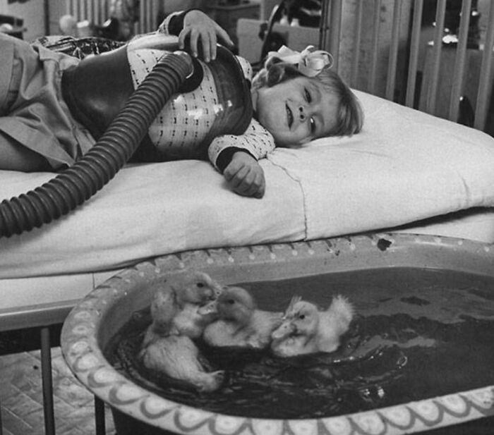 Niña recibiendo terapia médica con dispositivo respiratorio junto a patitos en agua, mostrando cuerpo y tratamiento médico.