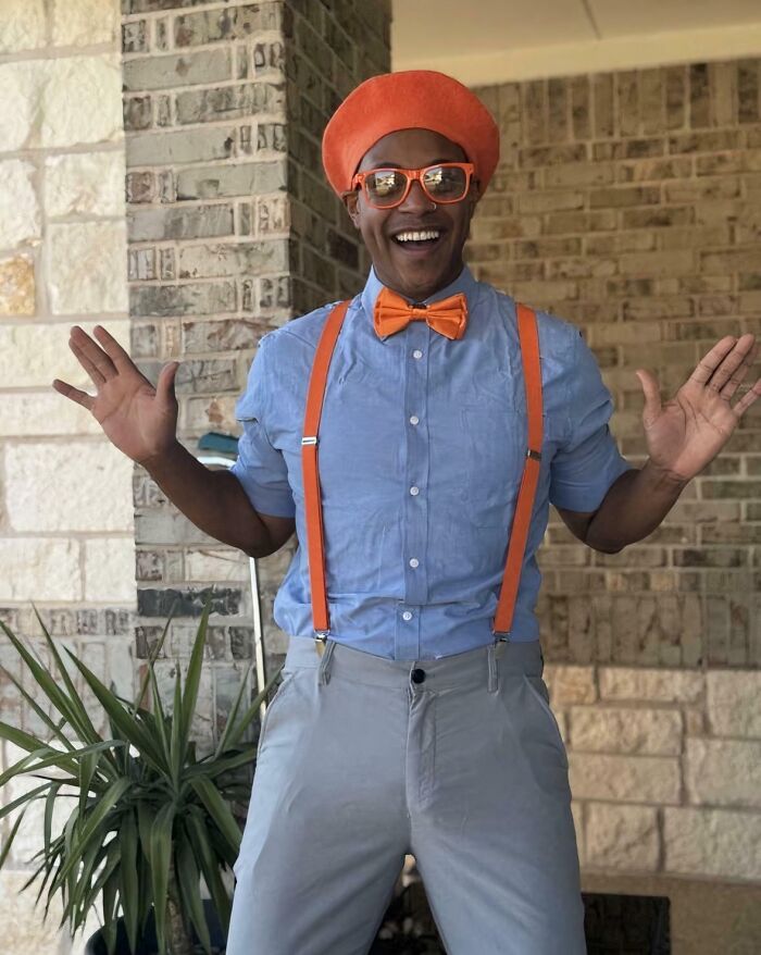 Padre con gafas, tirantes y moño naranja sonríe alegre mostrando divertida y cariñosa actitud parental.