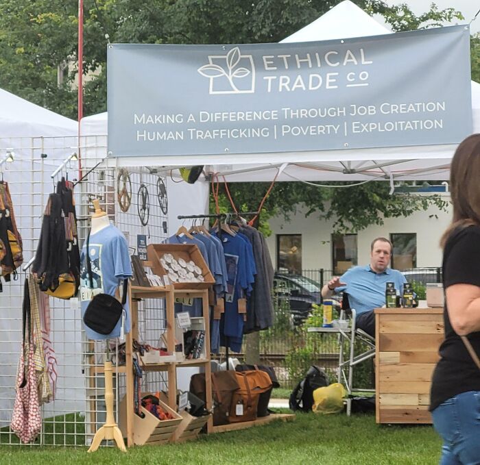 Puesto de venta de ropa y accesorios con cartel de Ethical Trade en feria al aire libre, mostrando divertidos fallos publicitarios.