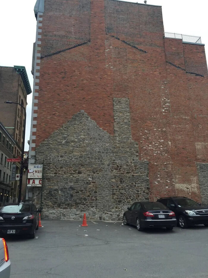 Edificio con muro de piedra antigua y ladrillos rojos en zona urbana con autos estacionados y conos naranjas visibles.