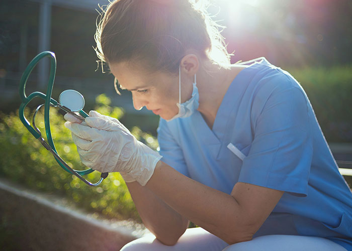 Médica en uniforme azul con guantes y cubrebocas, mirando un estetoscopio en un espacio exterior iluminado.