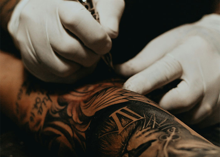 Manos con guantes realizando tatuaje en brazo con diseño complejo, ilustrando casos médicos horribles por retraso.