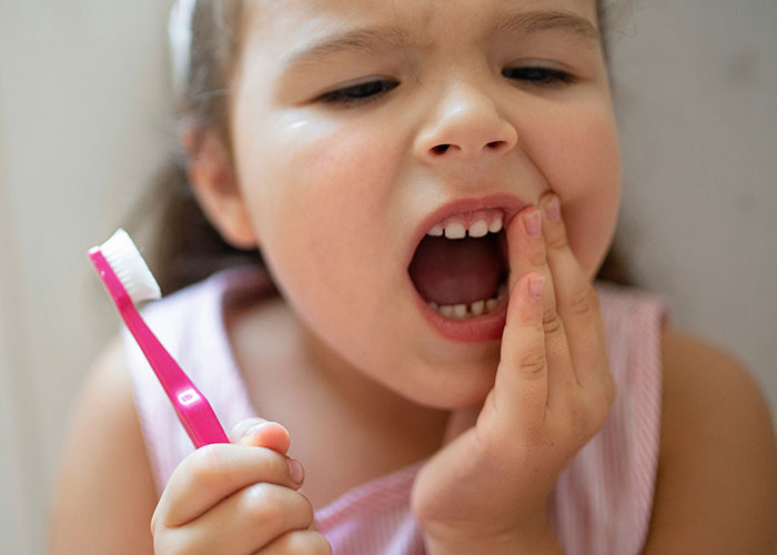 Niña con dolor dental sostiene un cepillo de dientes, reflejando casos horribles que médicos alertan deberían tratarse antes.
