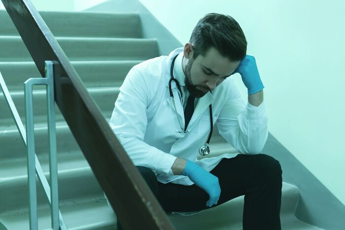 Médico con bata y guantes azules sentado en escaleras, mostrando señales de estrés tras escuchar algo impactante.