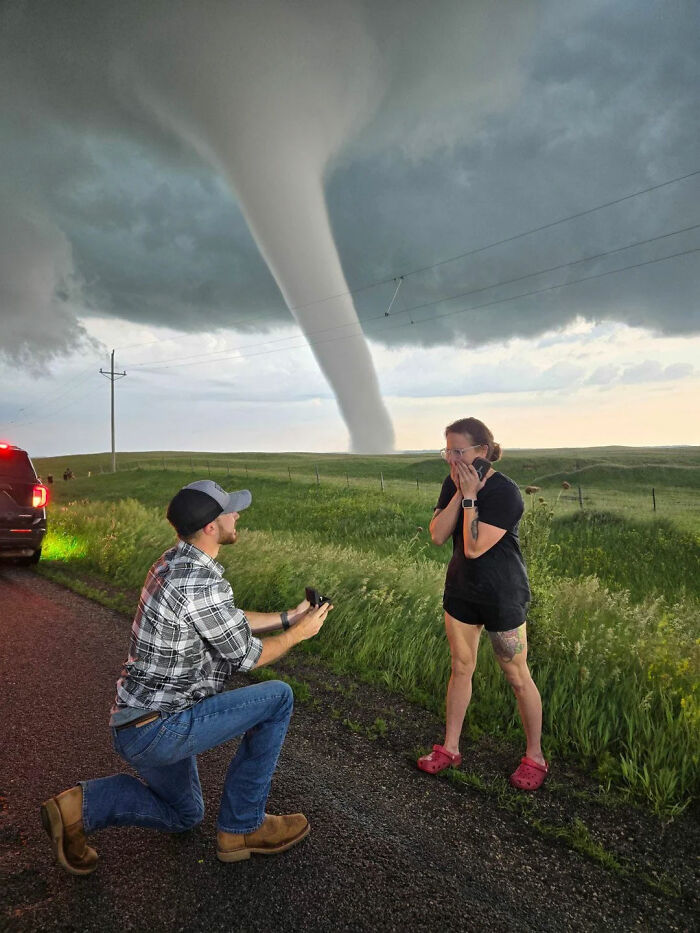 Hombre propone matrimonio cerca de un tornado enorme en campo verde con cielo nublado y mujer sorprendida.