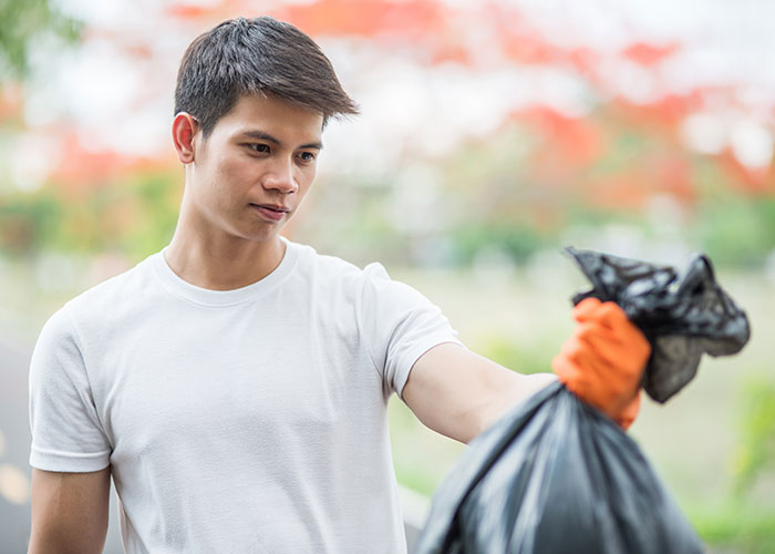 Joven con instinto de supervivencia sostiene una bolsa de basura negra usando guante naranja en un entorno exterior.