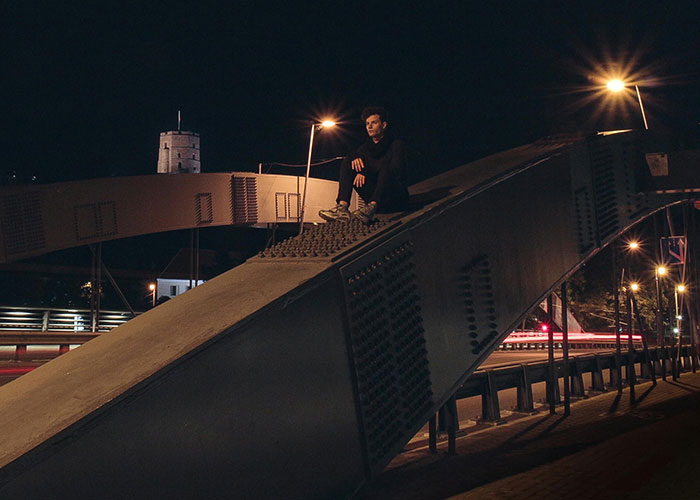 Joven sentado en estructura metálica de puente de noche, representando instinto de supervivencia en ambiente urbano.