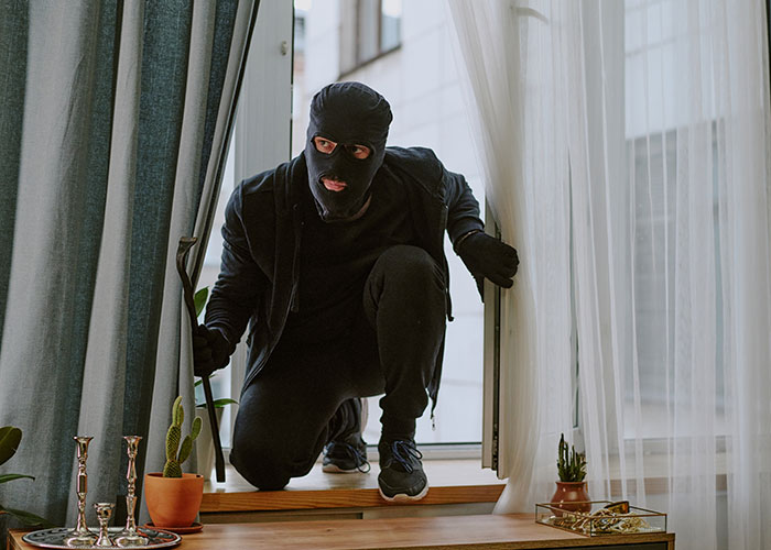Hombre con instinto de supervivencia entrando sigilosamente por ventana con ropa negra y pasamontañas en casa.
