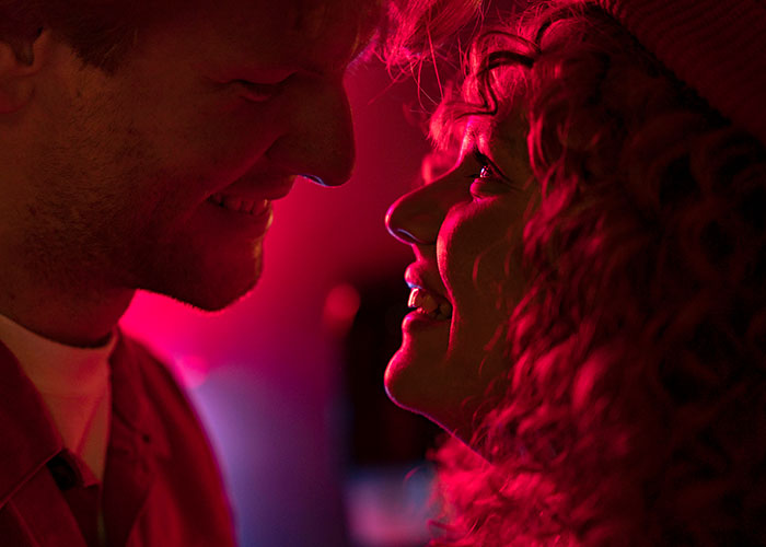 Pareja sonriendo y mirándose de cerca en ambiente con luz roja, mostrando instinto de supervivencia y conexión emocional.