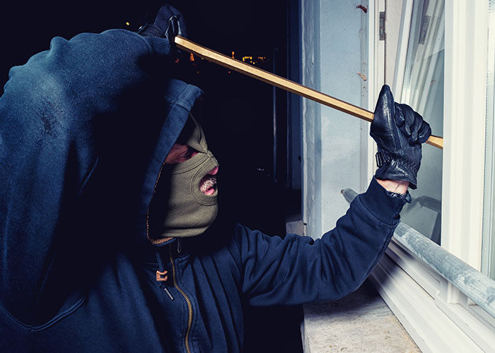Hombre con instinto de supervivencia intentando forzar una ventana por la noche con máscara y guantes negros.