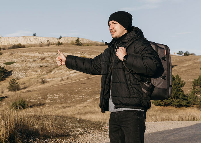 Hombre con instinto de supervivencia haciendo autoestop en paisaje abierto con mochila y ropa de invierno negra.