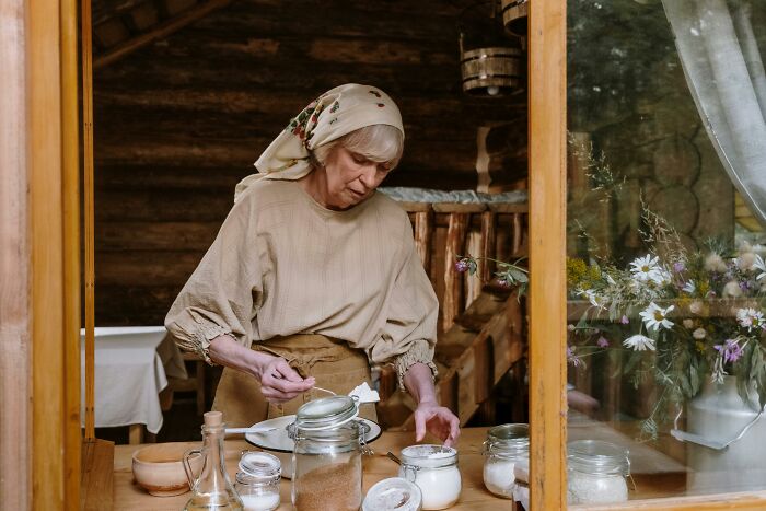 Mujer mayor en cocina rústica, preparando alimentos mientras reflexiona sobre secretos familiares desquiciados y deprimentes.