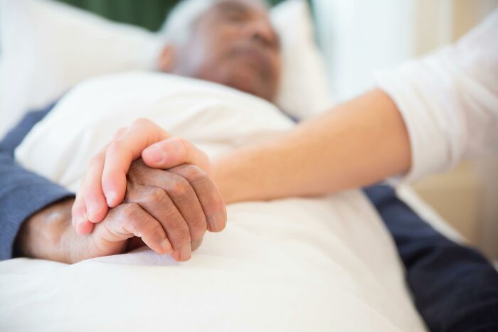 Mano de persona joven sosteniendo mano de adulto mayor en cama, mostrando conexión familiar y apoyo emocional.