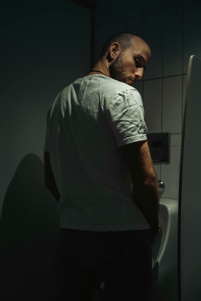 Hombre con camiseta blanca en baño oscuro, reflejando la incomodidad de escuchar accidentalmente peores cosas inolvidables.
