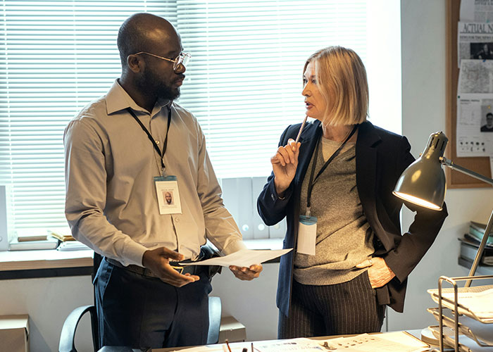 Dos investigadores privados discuten casos extraños mientras revisan documentos en una oficina iluminada con luz natural.