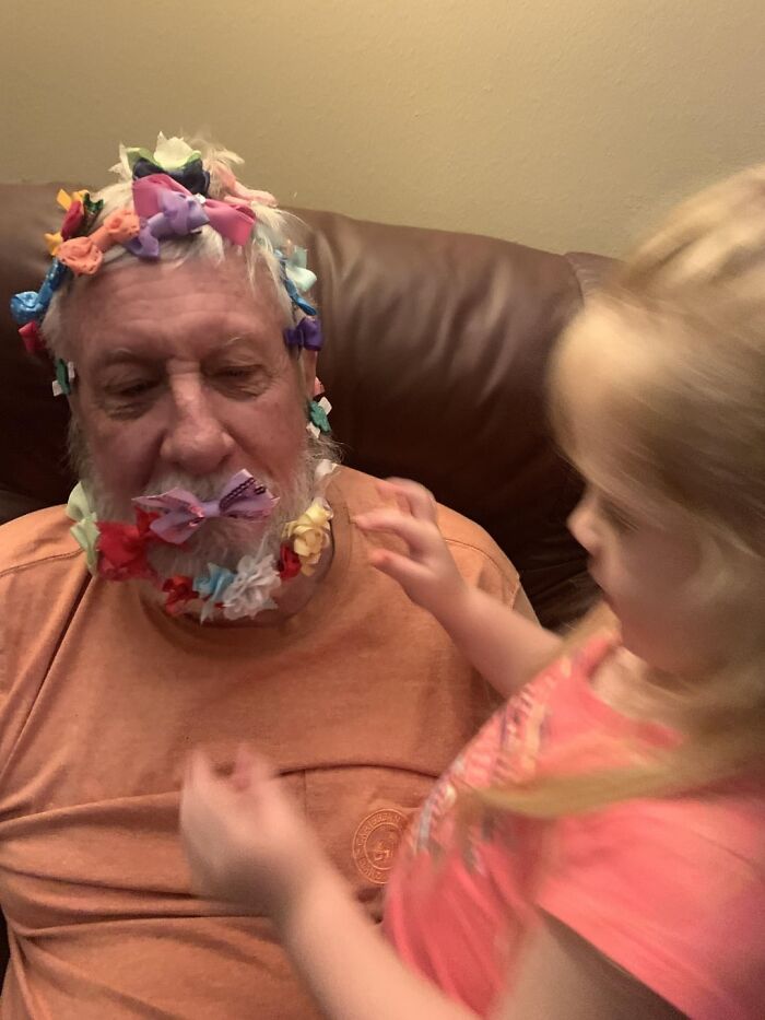 Hombre mayor con la barba y el cabello cubiertos de lazos de colores mientras una niña juega con ellos en casa.