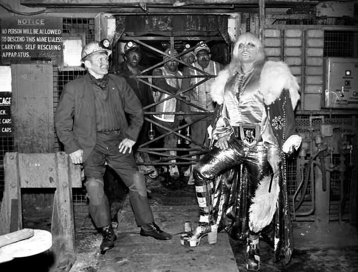 Fotografía histórica en negro y blanco mostrando mineros y una persona con vestimenta llamativa en una mina antigua.