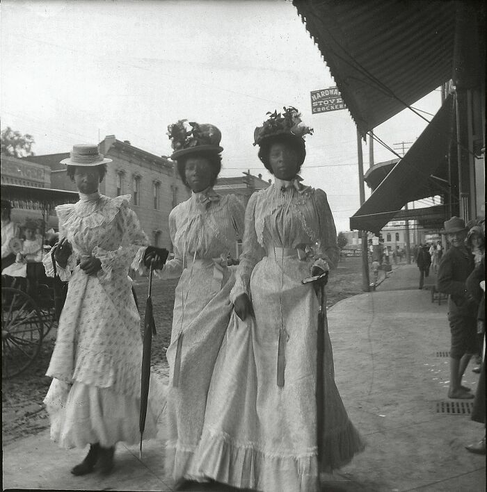 Tres mujeres vestidas con trajes antiguos y sombreros, en una calle con edificios y carrozas, foto histórica antigua.