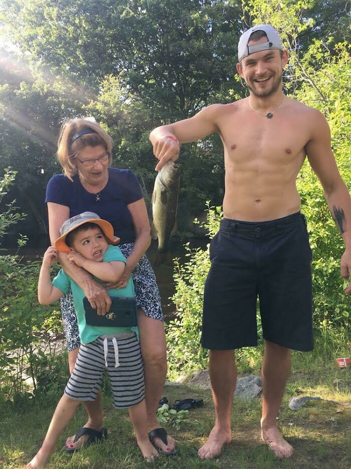 Hombre sonriente mostrando pez pescado en vacaciones de verano con niño y mujer mayor en un entorno natural divertido.